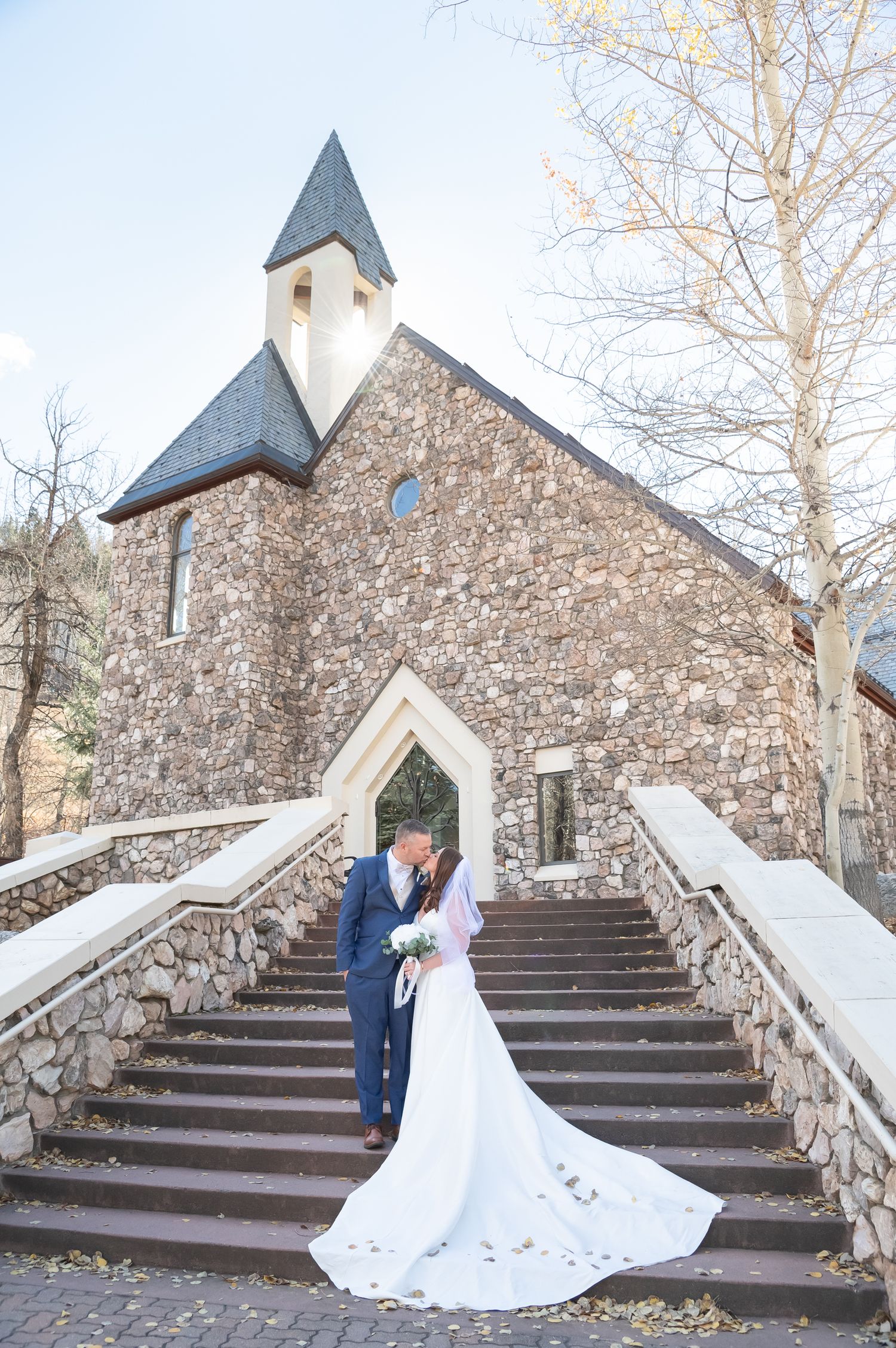 A romantic wedding couple poses on stone steps outside a rustic stone chapel with spires.