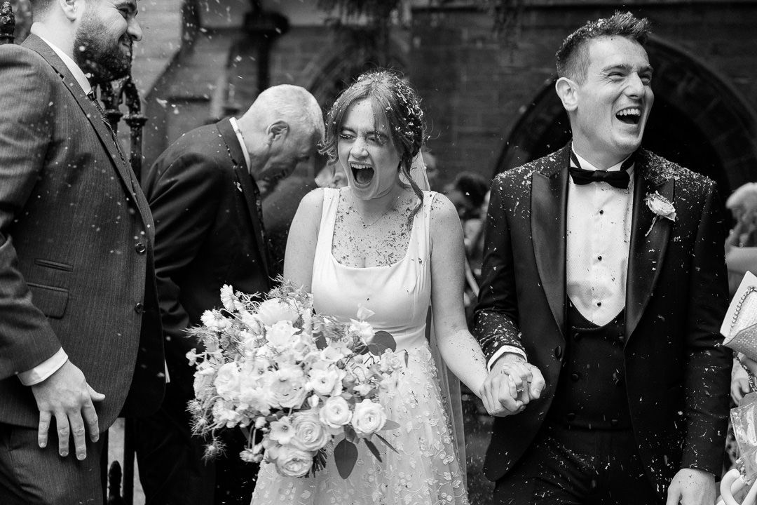 A bride and groom share candid moments while walking together on gravel outside a red brick building.