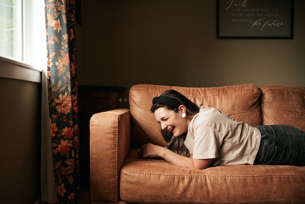 A young woman laughs while lying on a brown leather sofa near a window with floral curtains.