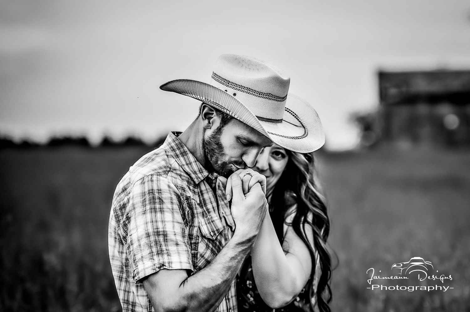 Romantic country couple embraces in rustic barn setting wearing cowboy hat and floral dress during engagement photoshoot.