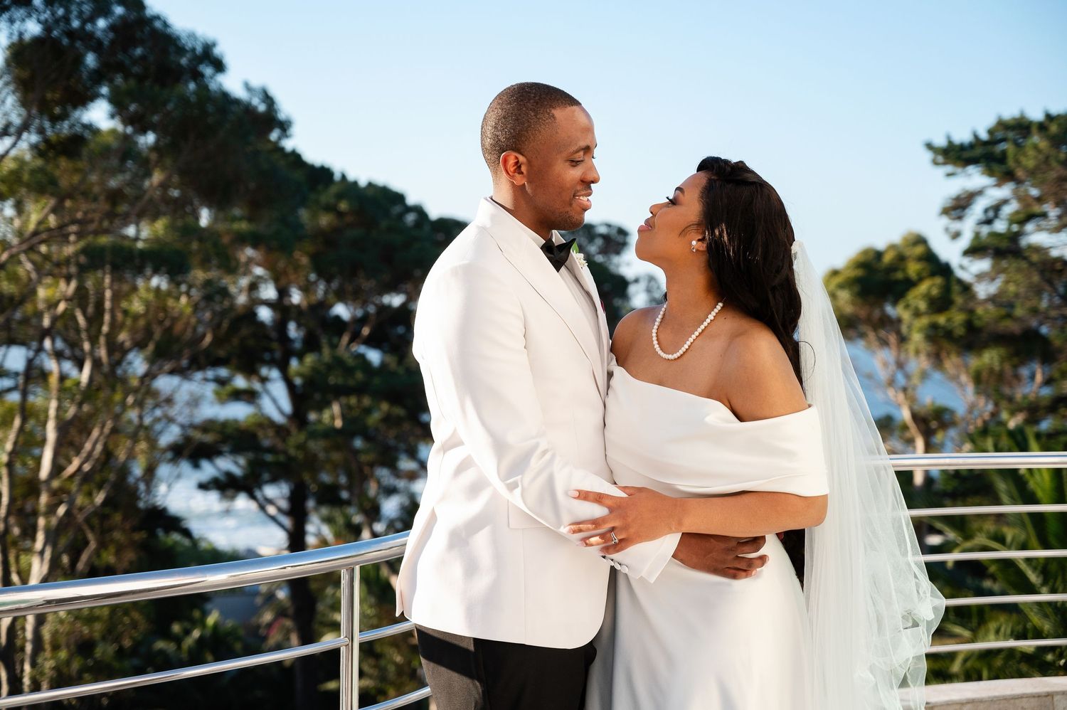 A couple in wedding attire embraces on a balcony overlooking a scenic coastal view during sunset.
