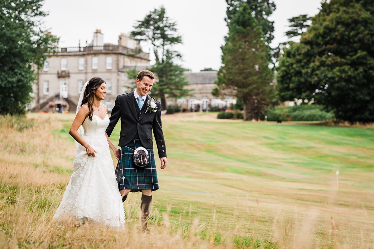 Bride in white dress, groom in kilt, laughing and holding hands at Dalmahoy Hotel Kirknewton with trees and golf course in background