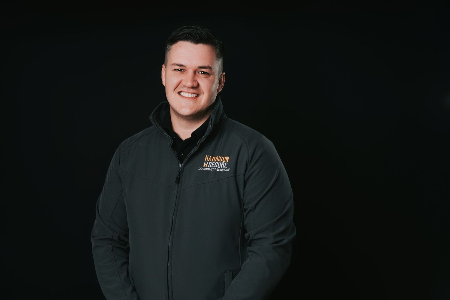 Professional headshot against dark background showing person wearing dark green zip-up jacket with embroidered logo.