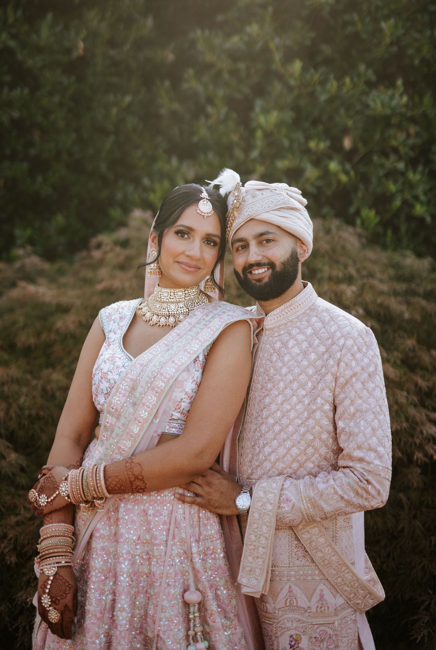 A couple in matching light pink and white traditional Indian wedding attire pose together in a garden setting.