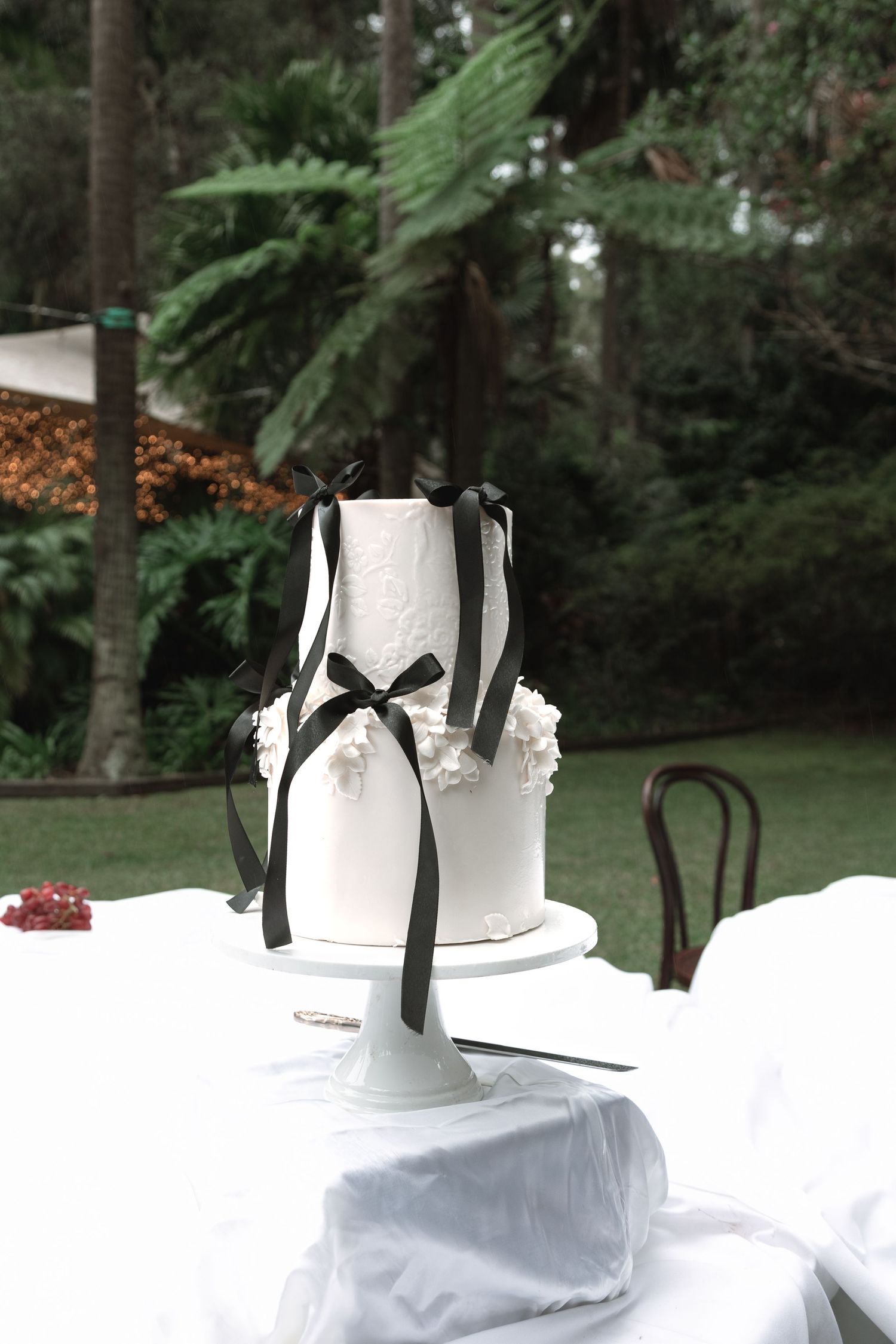 White ceramic vases with black ribbons arranged on white tablecloths at an outdoor garden event setup.