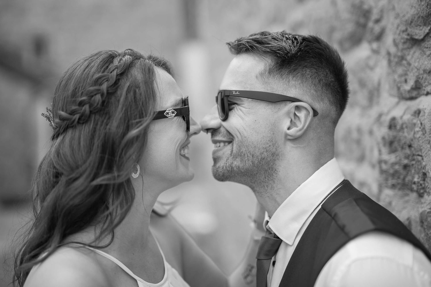 A romantic black and white portrait of a couple touching noses against a stone wall.