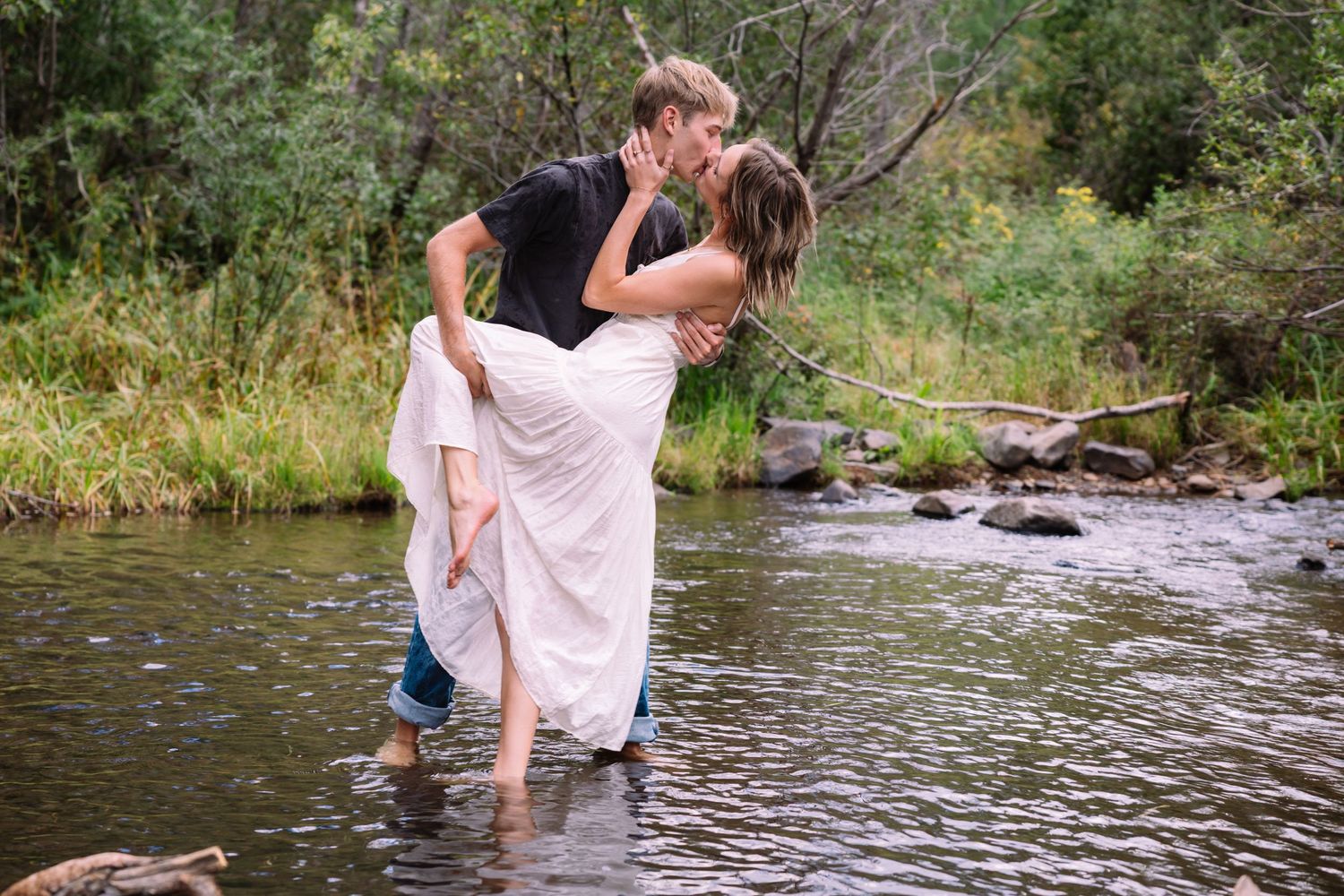 Two people play and splash in a shallow stream surrounded by green trees and vegetation in a natural setting.