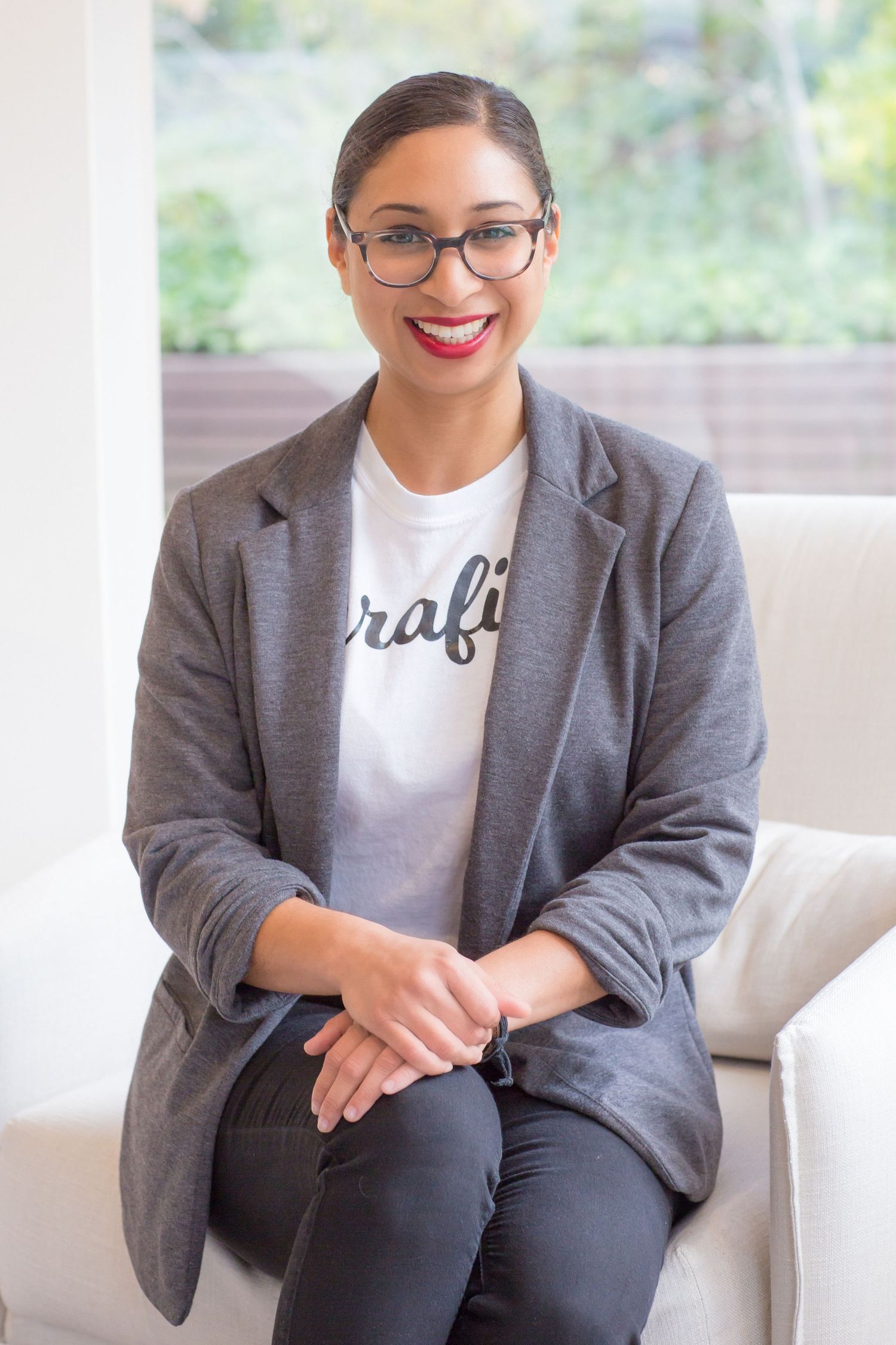 Professional business portrait of a person wearing glasses, a gray blazer and white t-shirt sitting in an office setting.