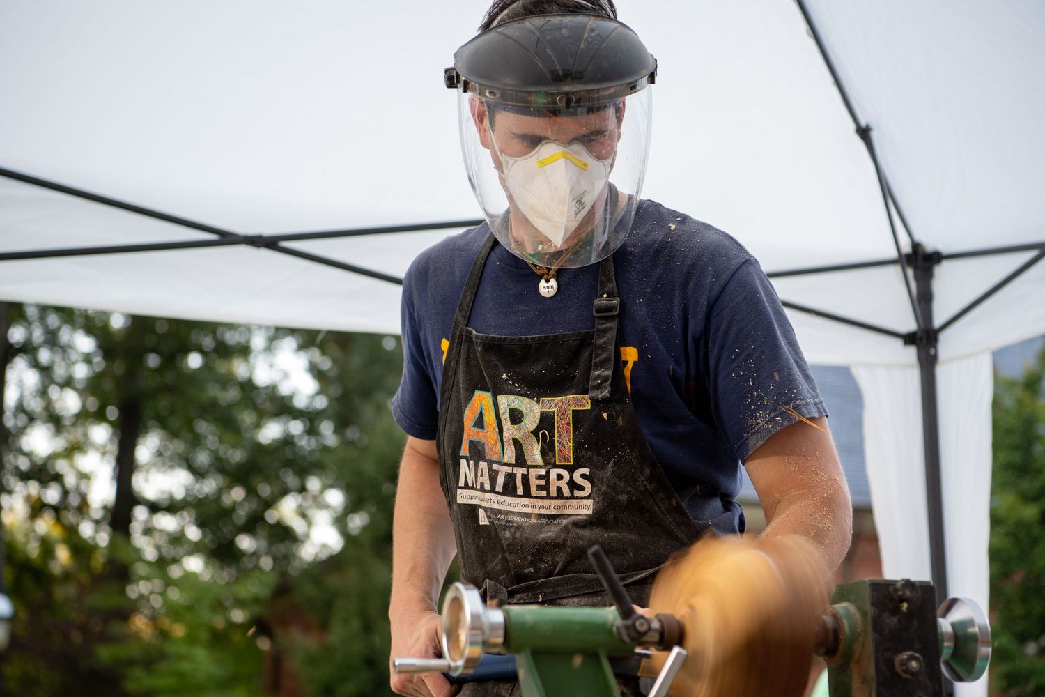 An artisan in a protective mask and dark shirt skillfully works with small metal objects at an outdoor market stall.