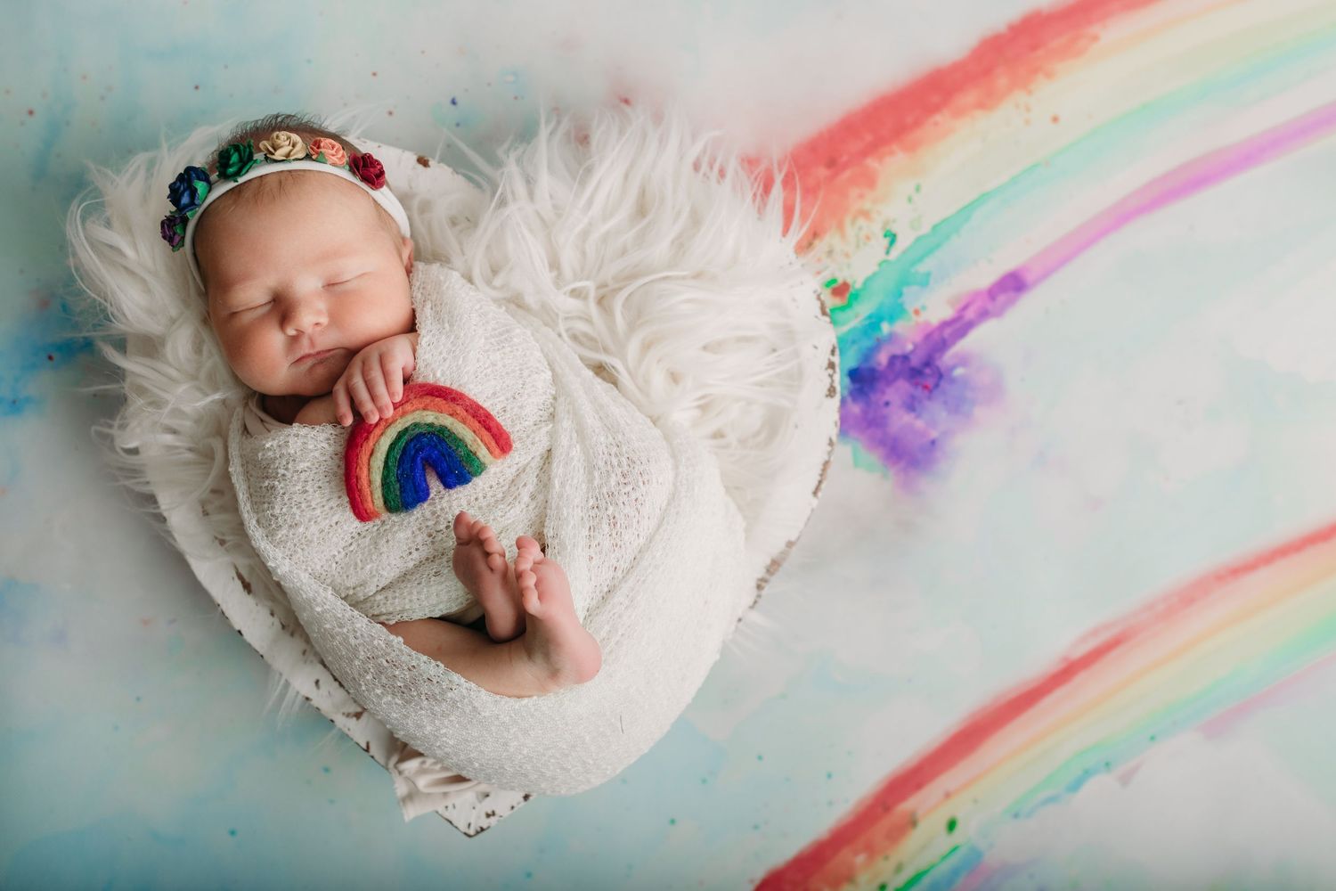 Sleeping newborn on white fur with rainbow decorations in dreamy pastel composition.