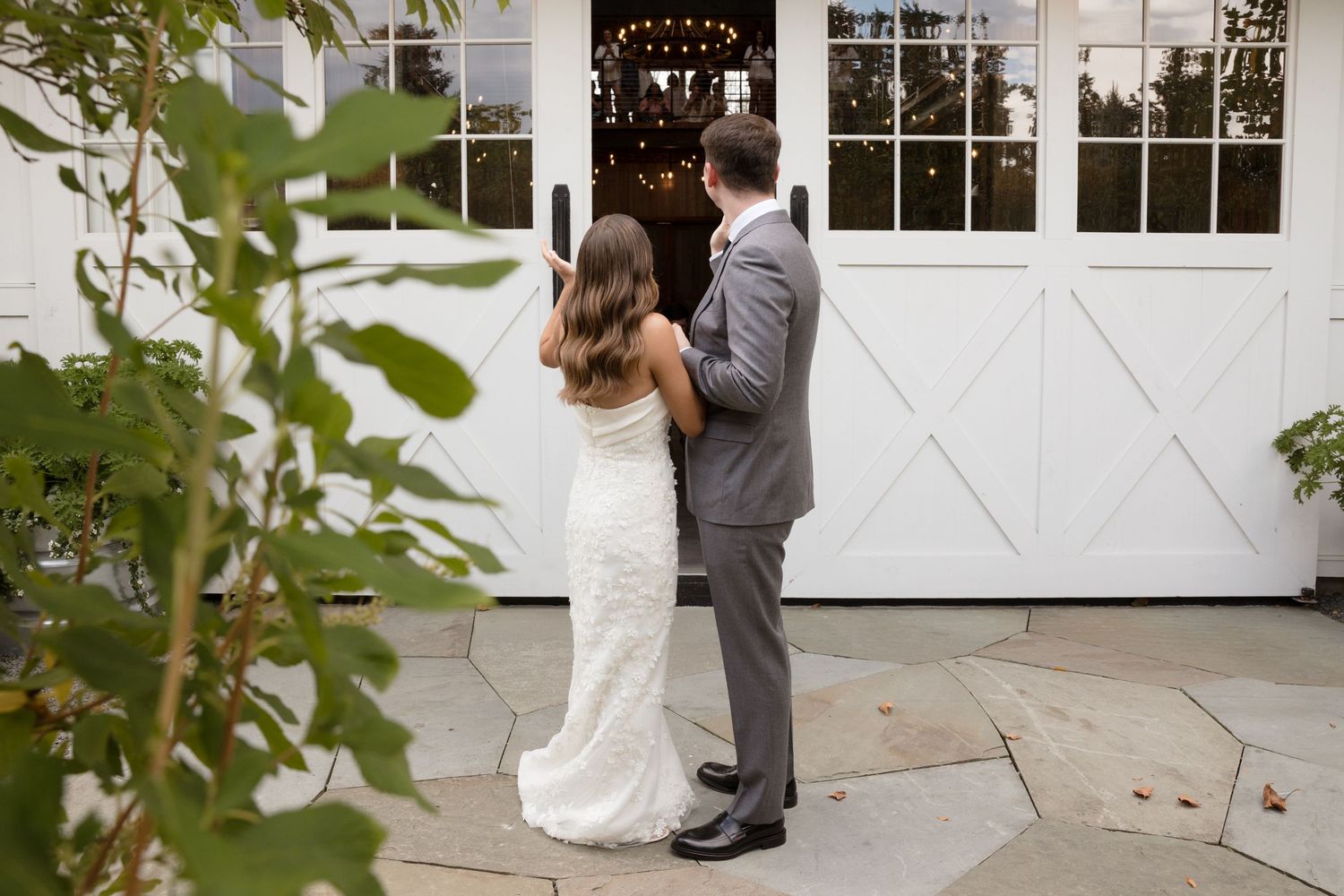 Wedding couple stands in front of a white building's entrance flanked by lush green foliage.