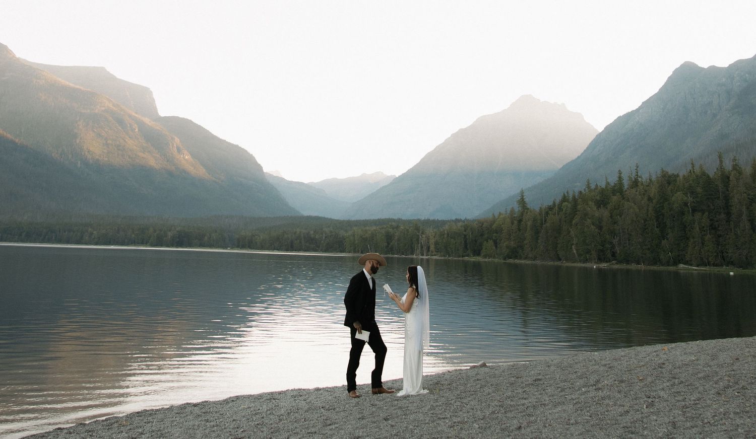 A couple stands in silhouette by a tranquil mountain lake at Glacier National Park during sunset.