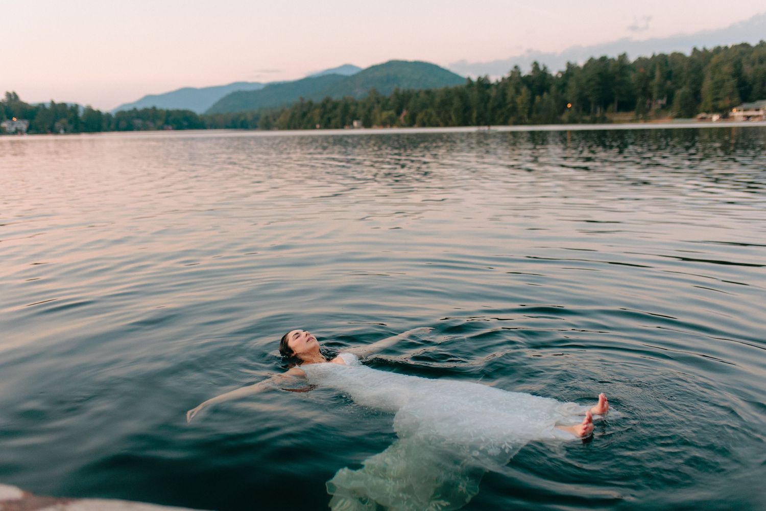 Someone floating on their back in a serene mountain lake during sunset with rippling water and forested shoreline.