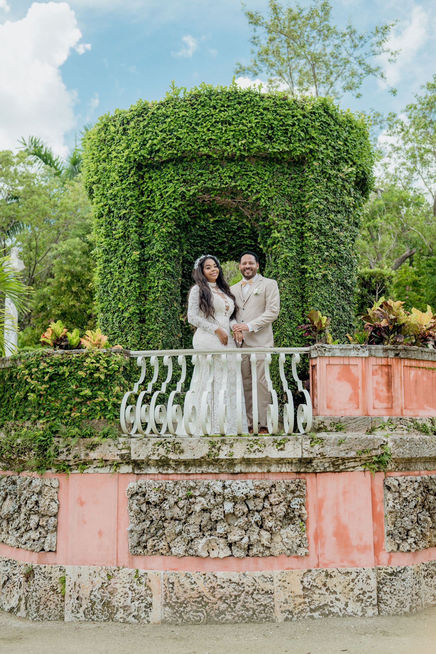 A joyful couple shares romantic moments in front of a lush green hedge and coral-colored stone wall in an outdoor garden setting.