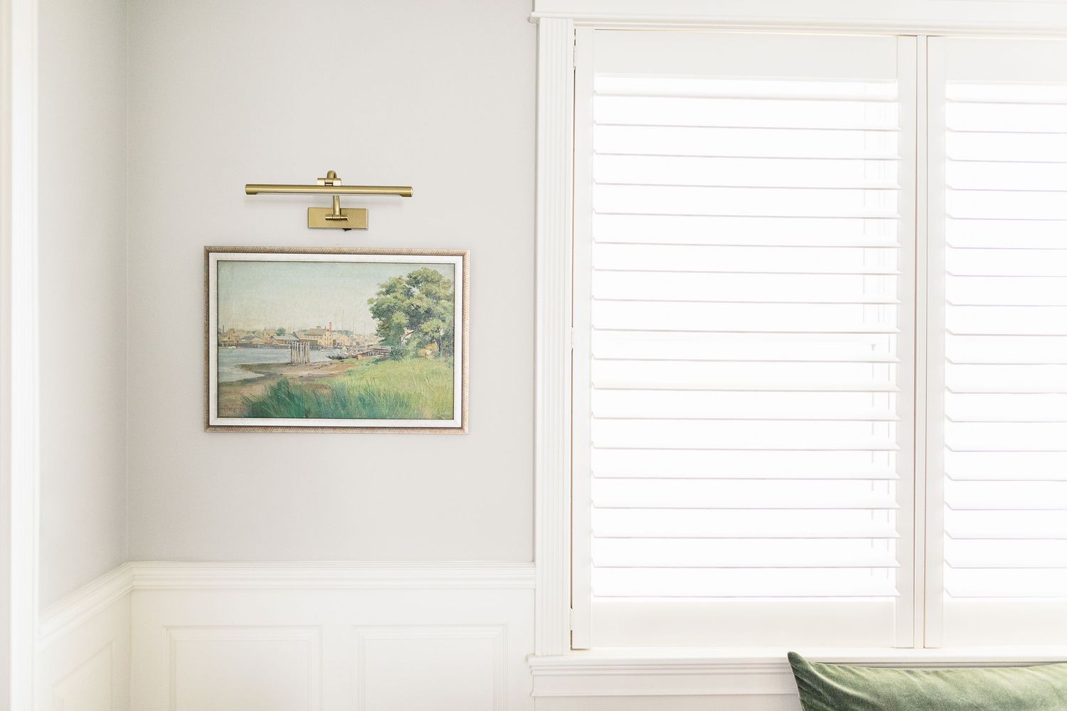 A bright white bedroom with a window, cream colored bed, and framed landscape artwork on the wall.