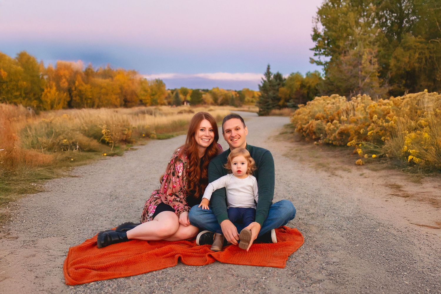 A family of 3 mom, dad and toddler girl sit on a blanket on a gravel path winds through autumn trees with golden foliage.