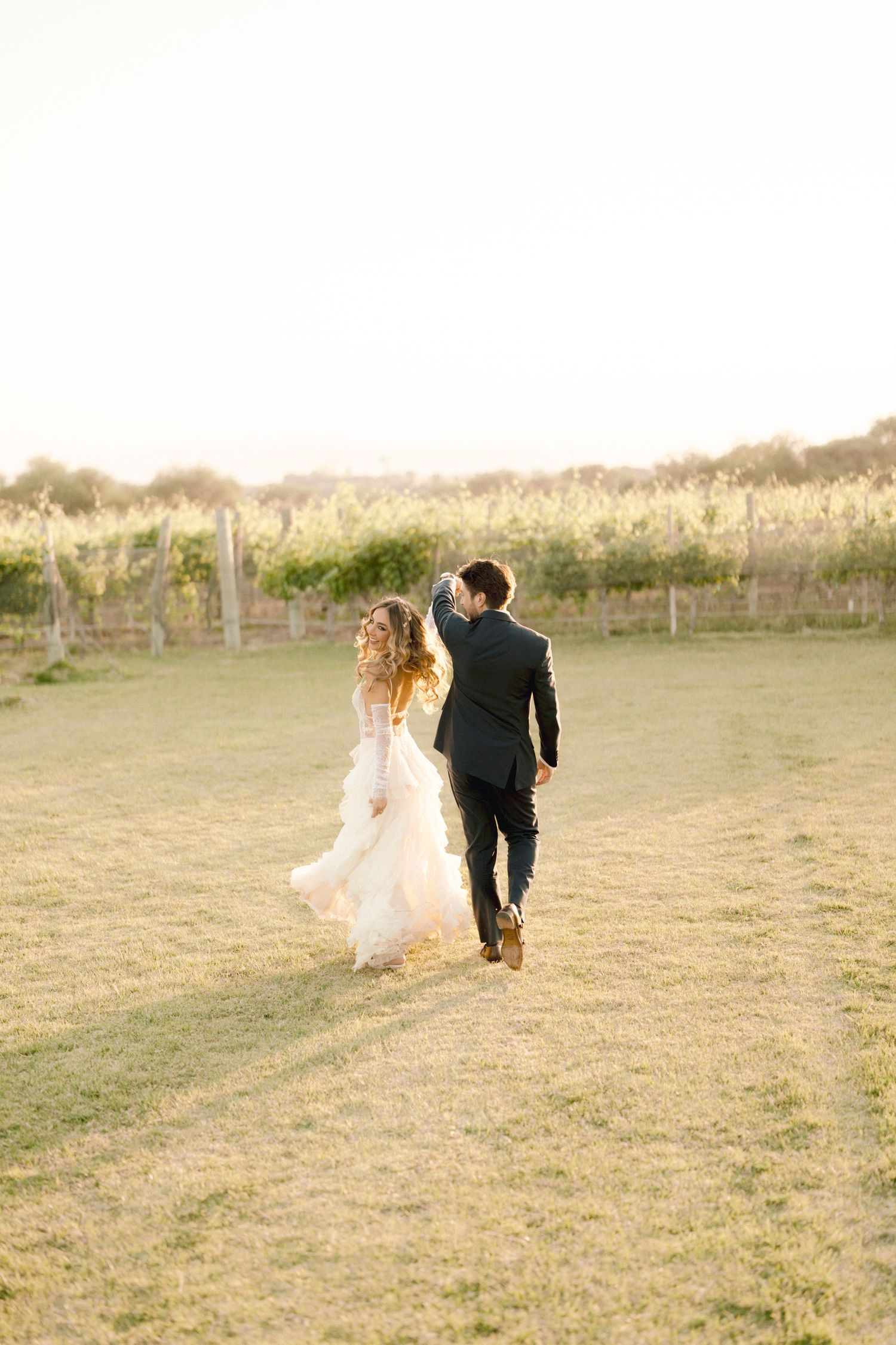 A couple runs playfully through a grassy field at sunset, with the bride's dress and hair flowing in the wind.