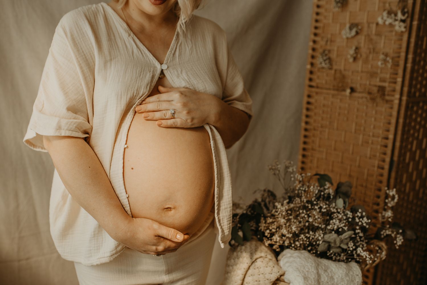 Woman in white shirt holding her pregnant belly while sitting in a warm-lit room with bamboo wall decor.