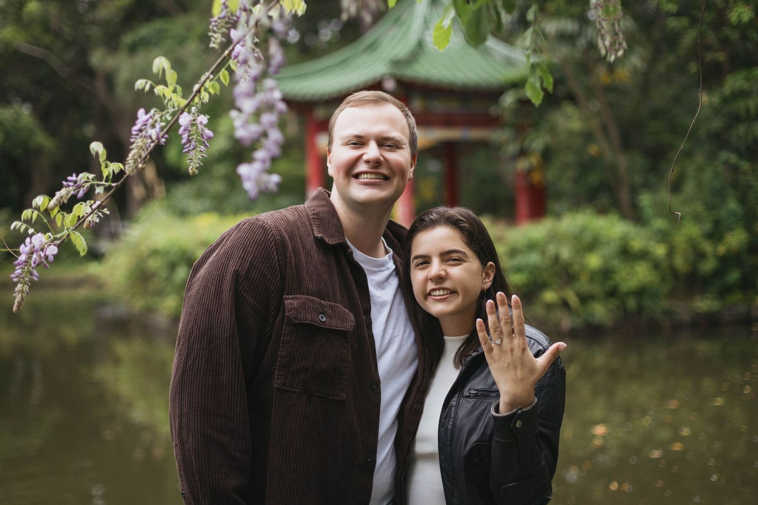 Happy couple posing near wisteria blooms with a red pagoda in a lush garden after engagement.