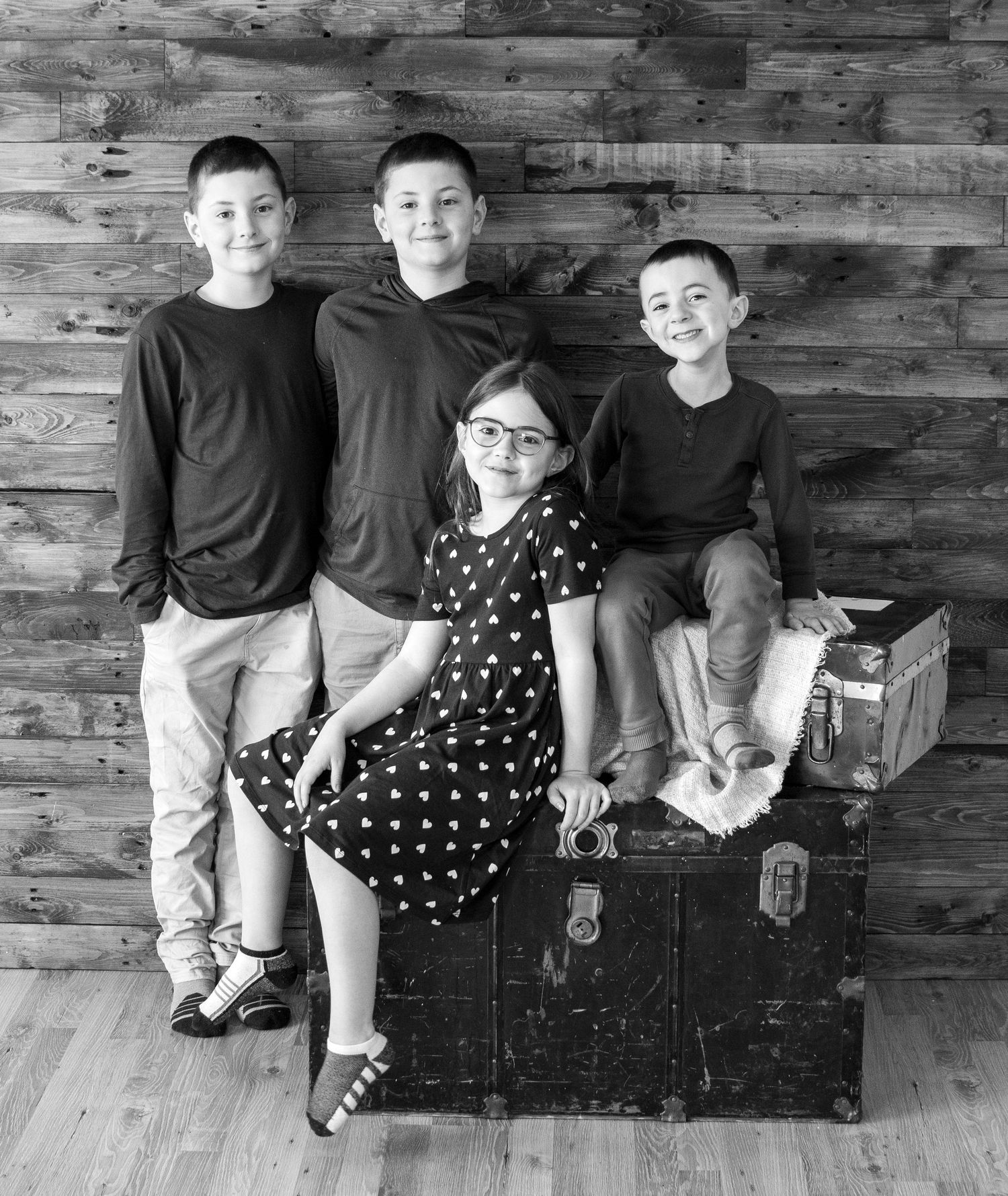 Black and white portrait of four siblings posed against a rustic wooden wall with the youngest sitting on a vintage trunk.