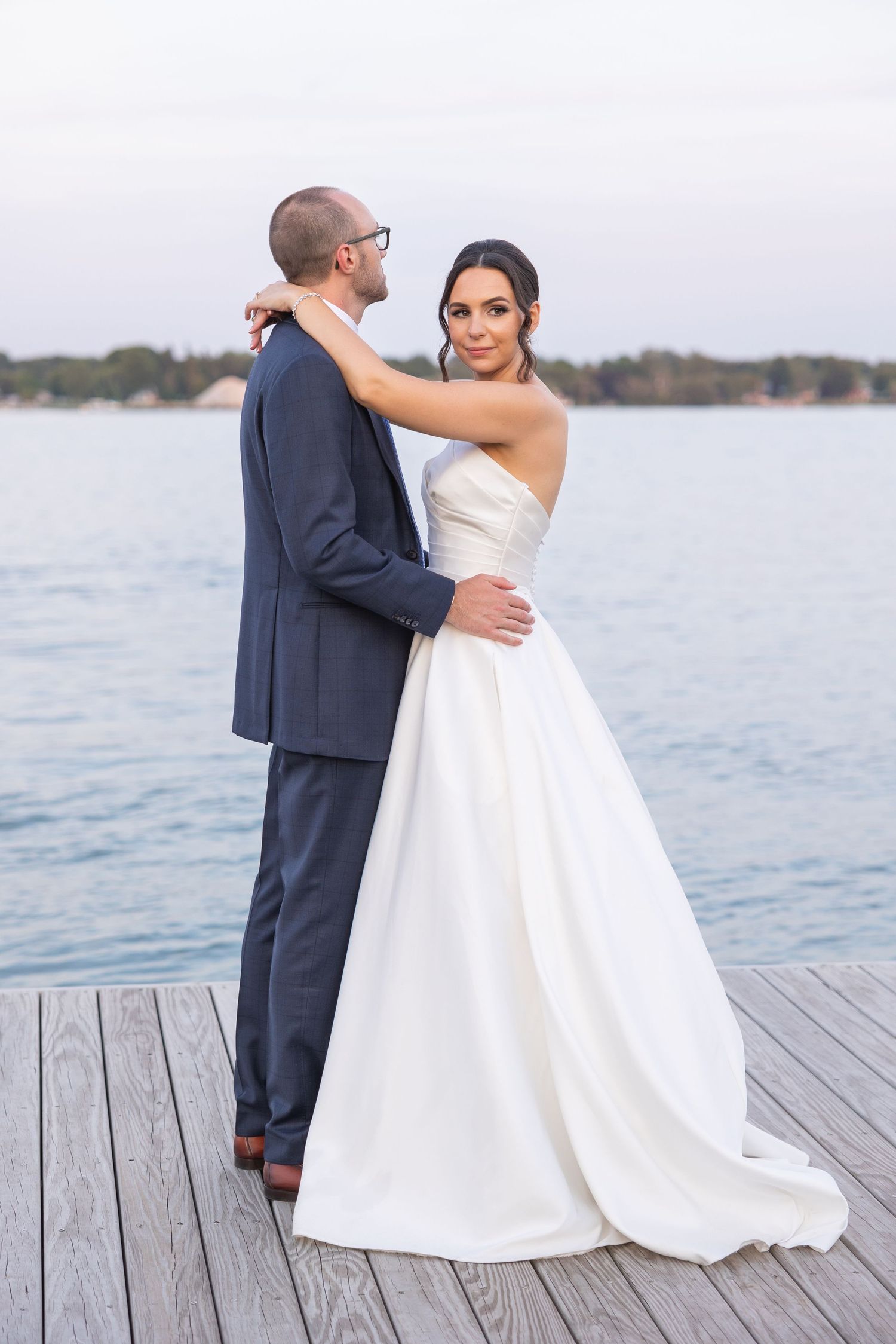 A bride in a strapless white gown and groom in navy suit embrace on a wooden dock by the water at sunset.