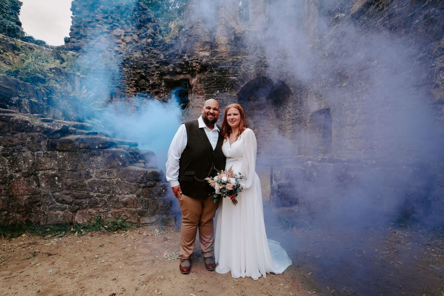 Wedding couple poses among lush greenery and white floral arrangements outdoors.