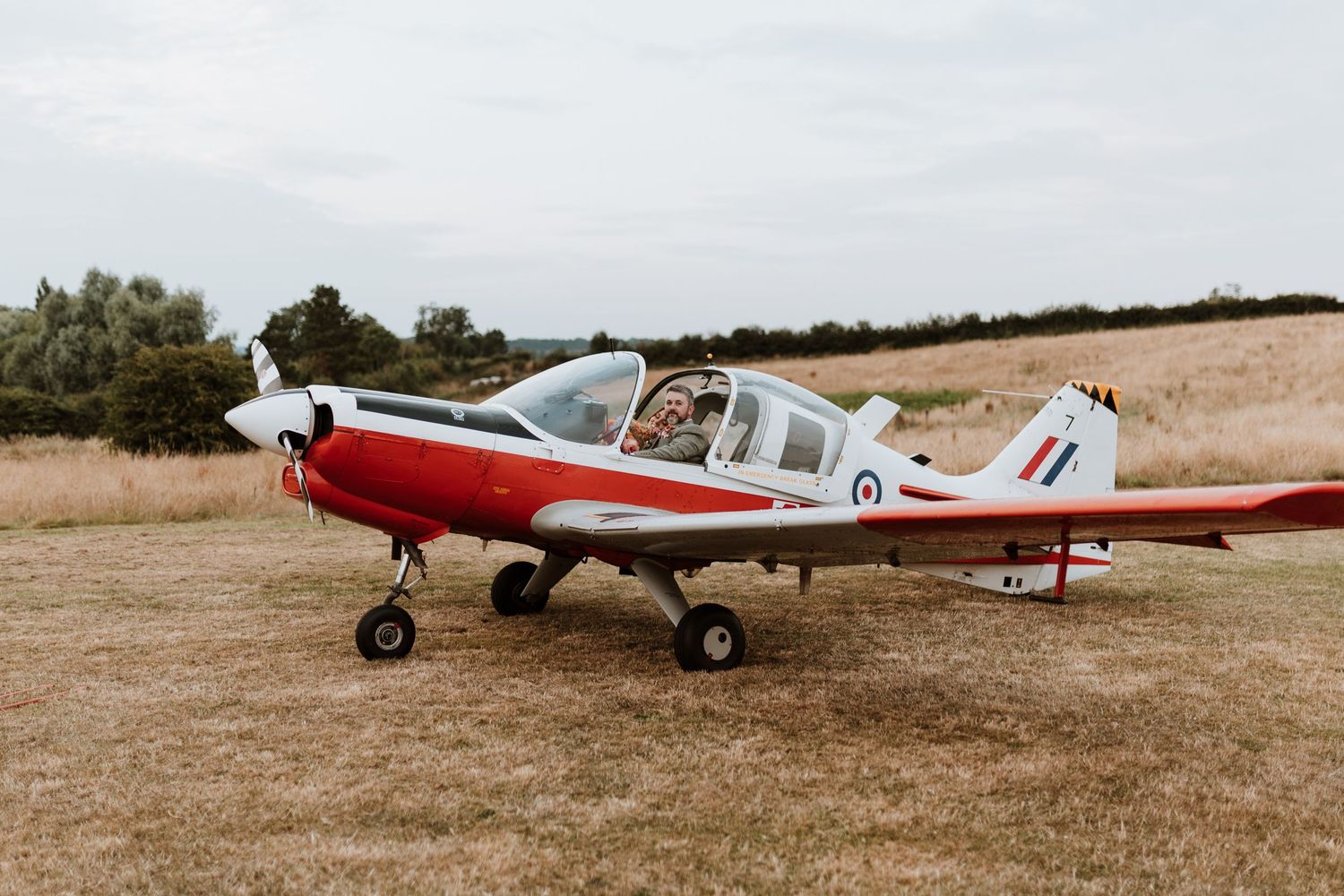A red and white small aircraft parked on a grass airfield with mountains visible in the background.