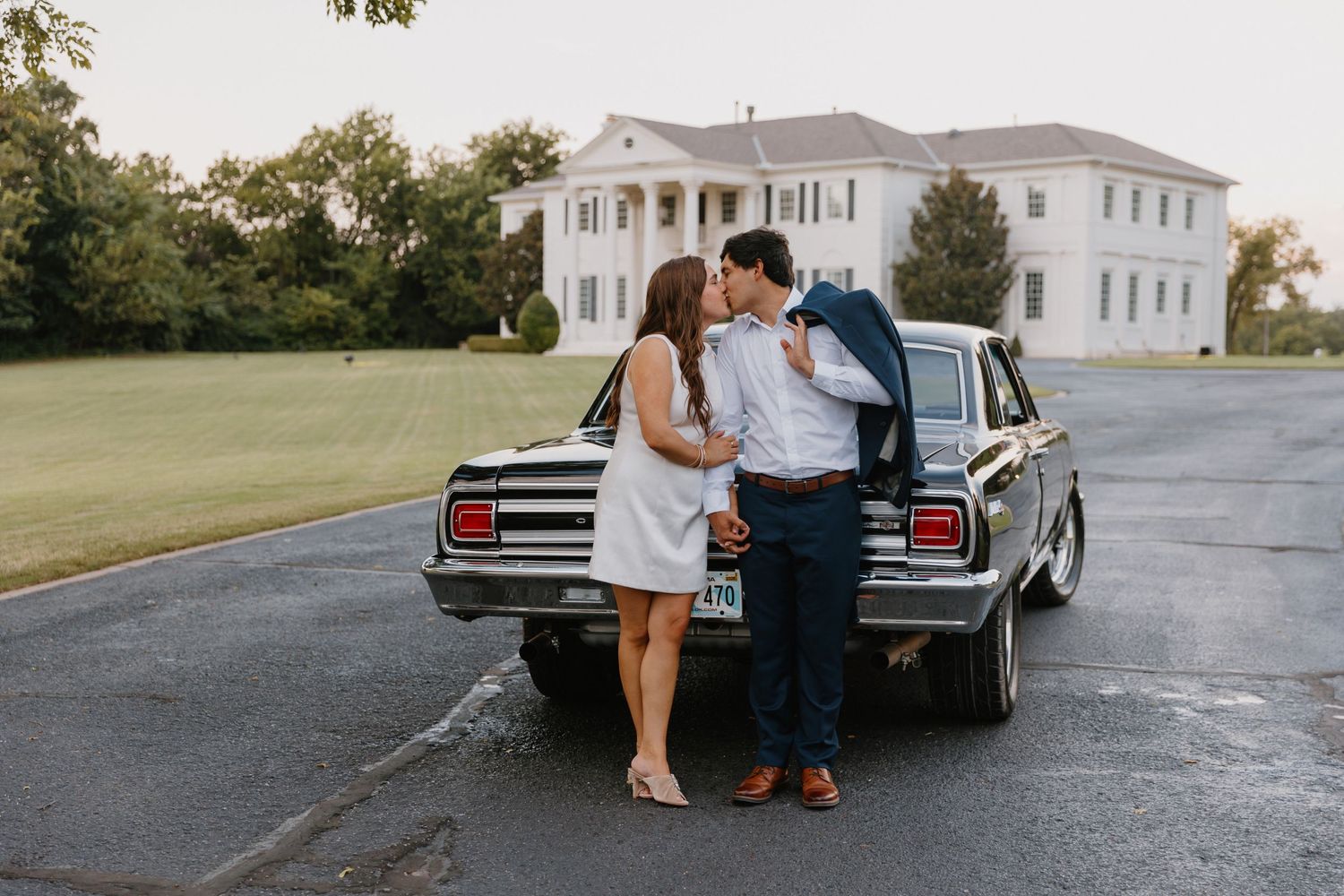 A couple sits on the back of a vintage car parked in front of a white colonial mansion.