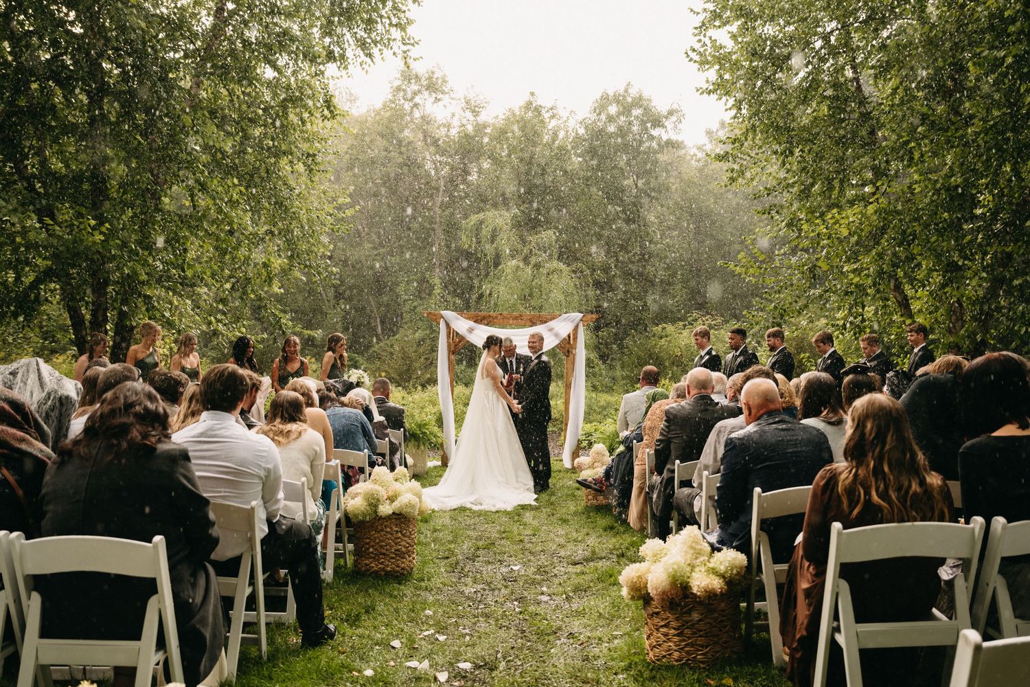 Outdoor wedding ceremony in a lush garden setting with guests seated on white chairs beneath tall trees.