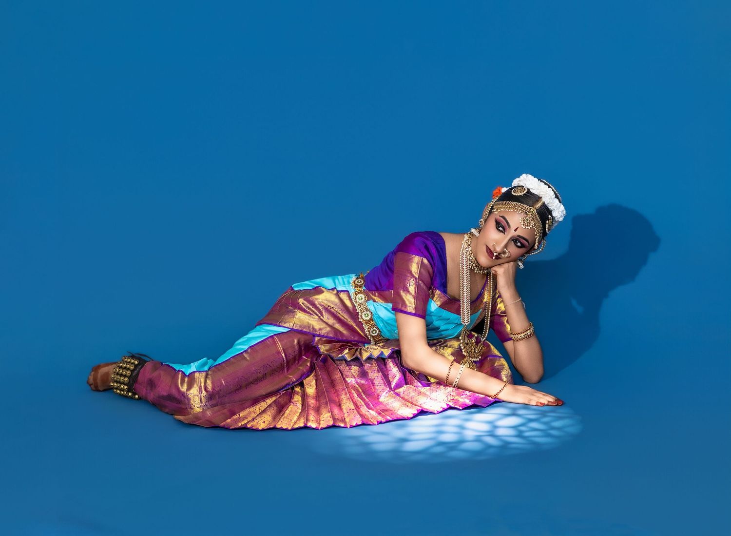 A person in a shimmering purple and gold traditional Indian dance costume reclines on a bright blue background.