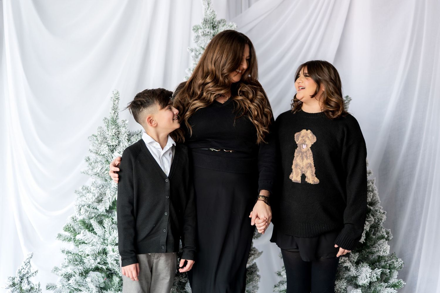 Mother and two children in black outfits holding hands in front of white drape with snowy trees.