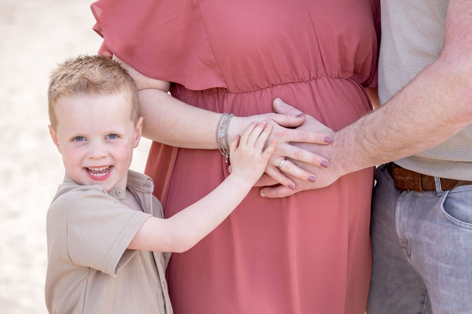 jongetje (4) samen met vader en moeder. Lachend richting de camera, waarbij hij en zijn ouders hun hand op de zwangere buik van mama leggen.