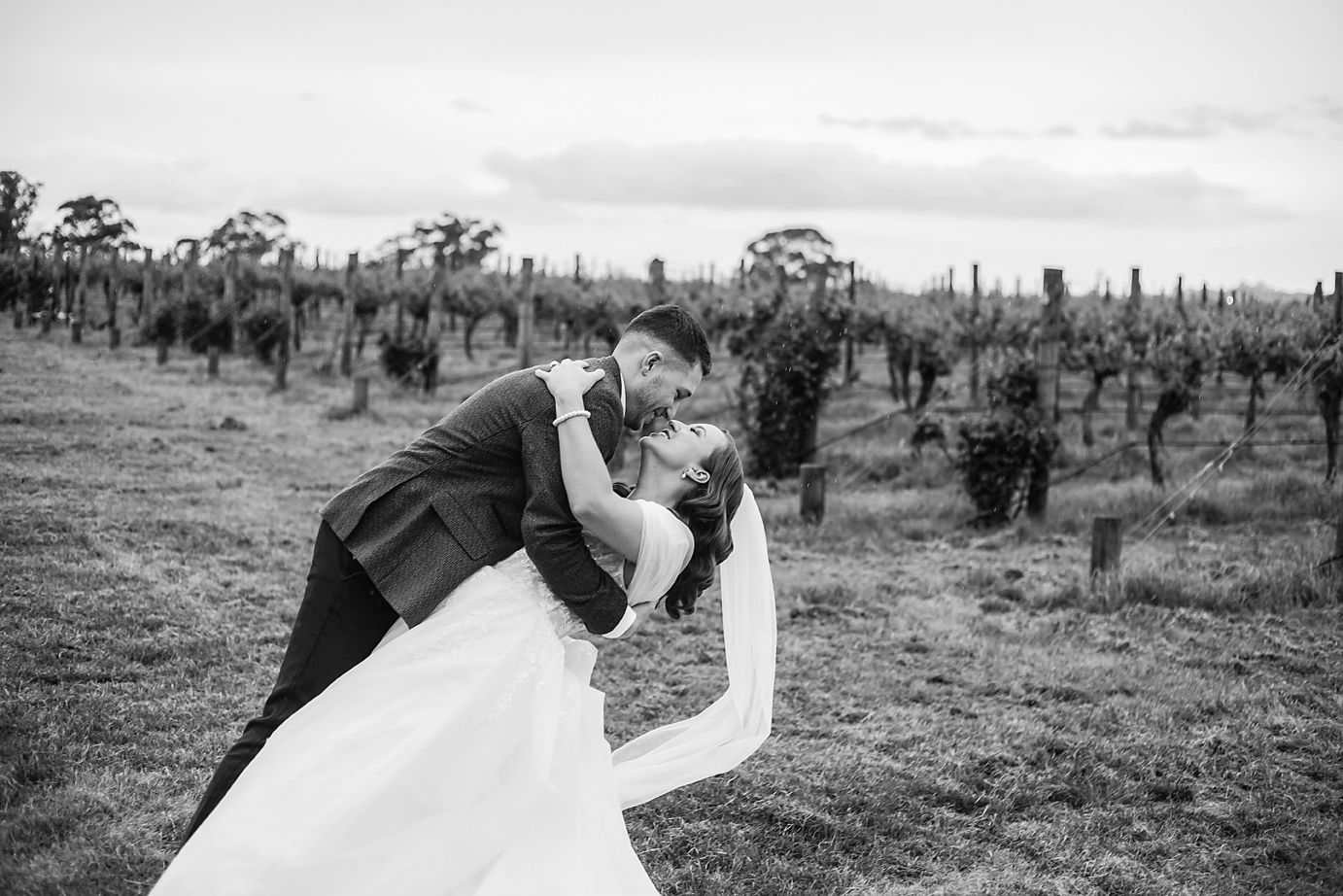 A couple in wedding attire dances together in a scenic vineyard setting at sunset.