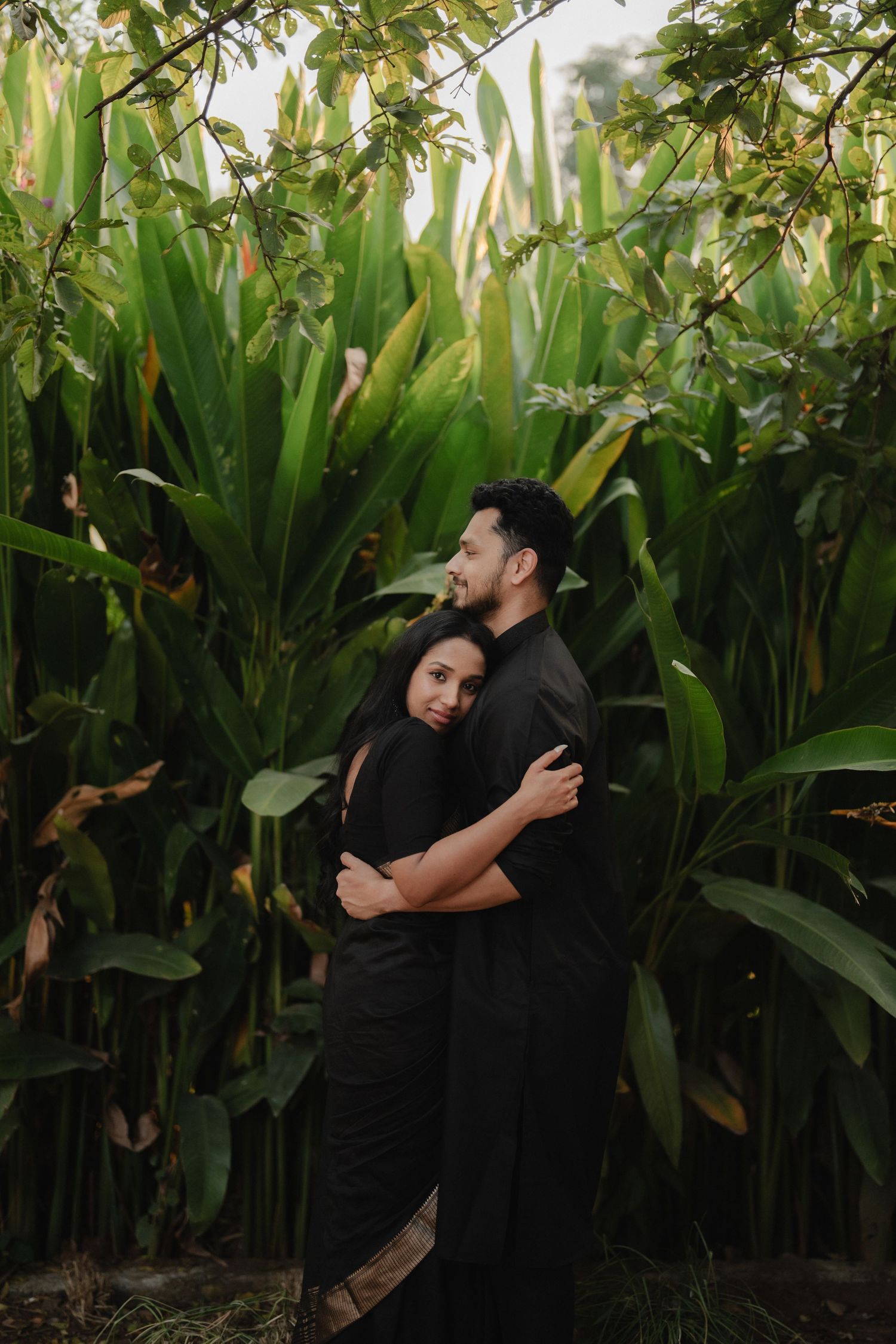 Two figures embrace in an intimate moment surrounded by tall green plants and foliage in a garden setting.