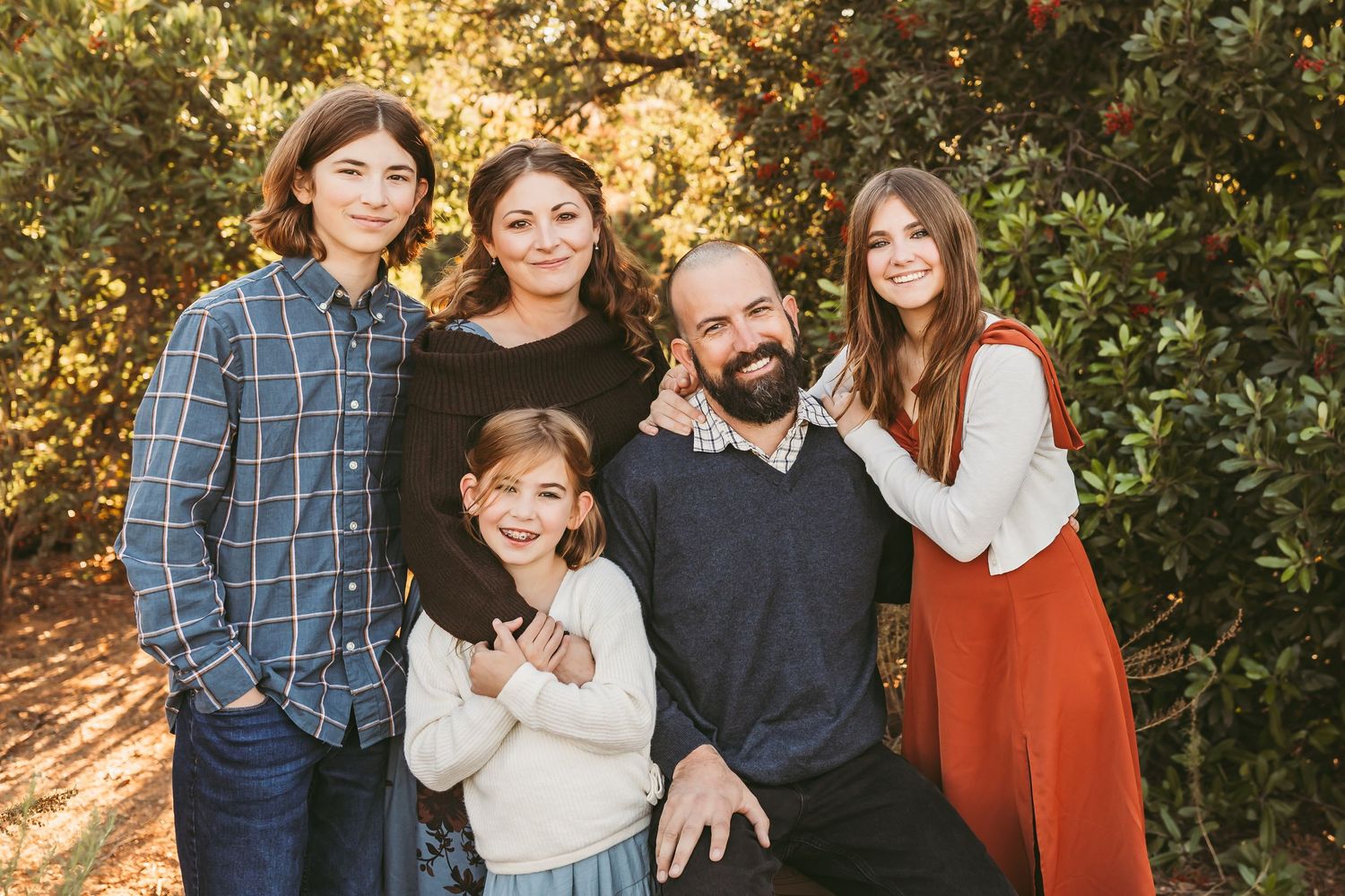 A family group poses together outdoors in coordinating autumn outfits against a sunlit natural background.
