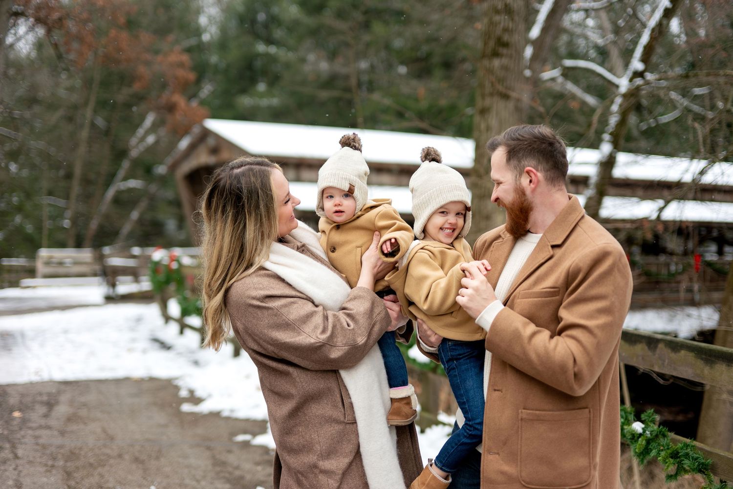 Family of four in matching tan coats enjoys snowy winter day near decorated covered bridge.