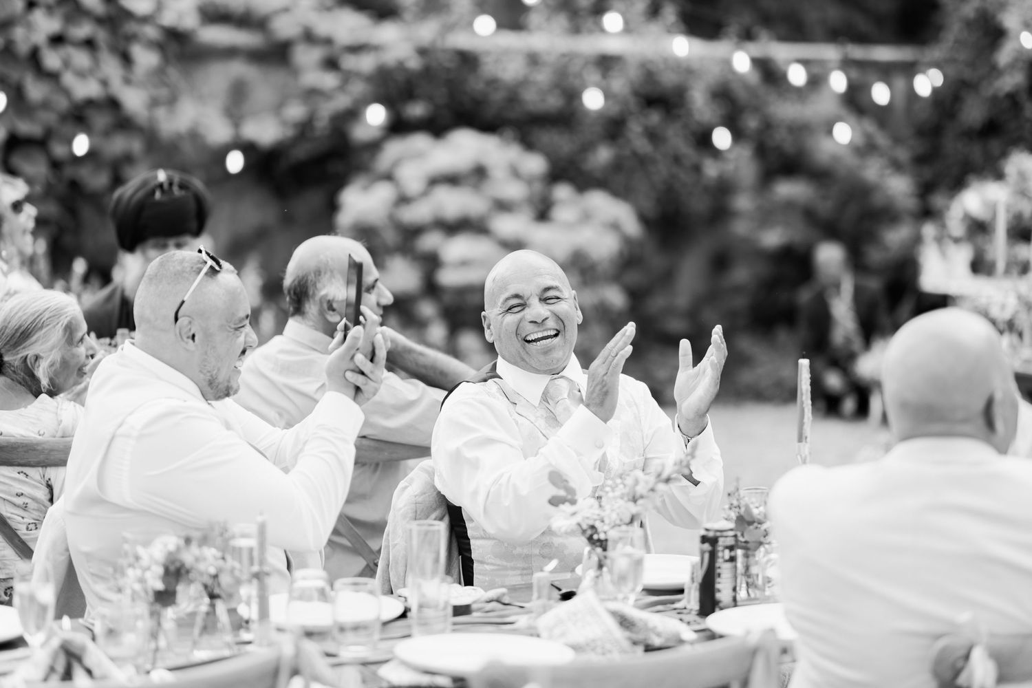 Black and white photograph showing a large outdoor wedding reception with guests seated at long tables in a garden setting.