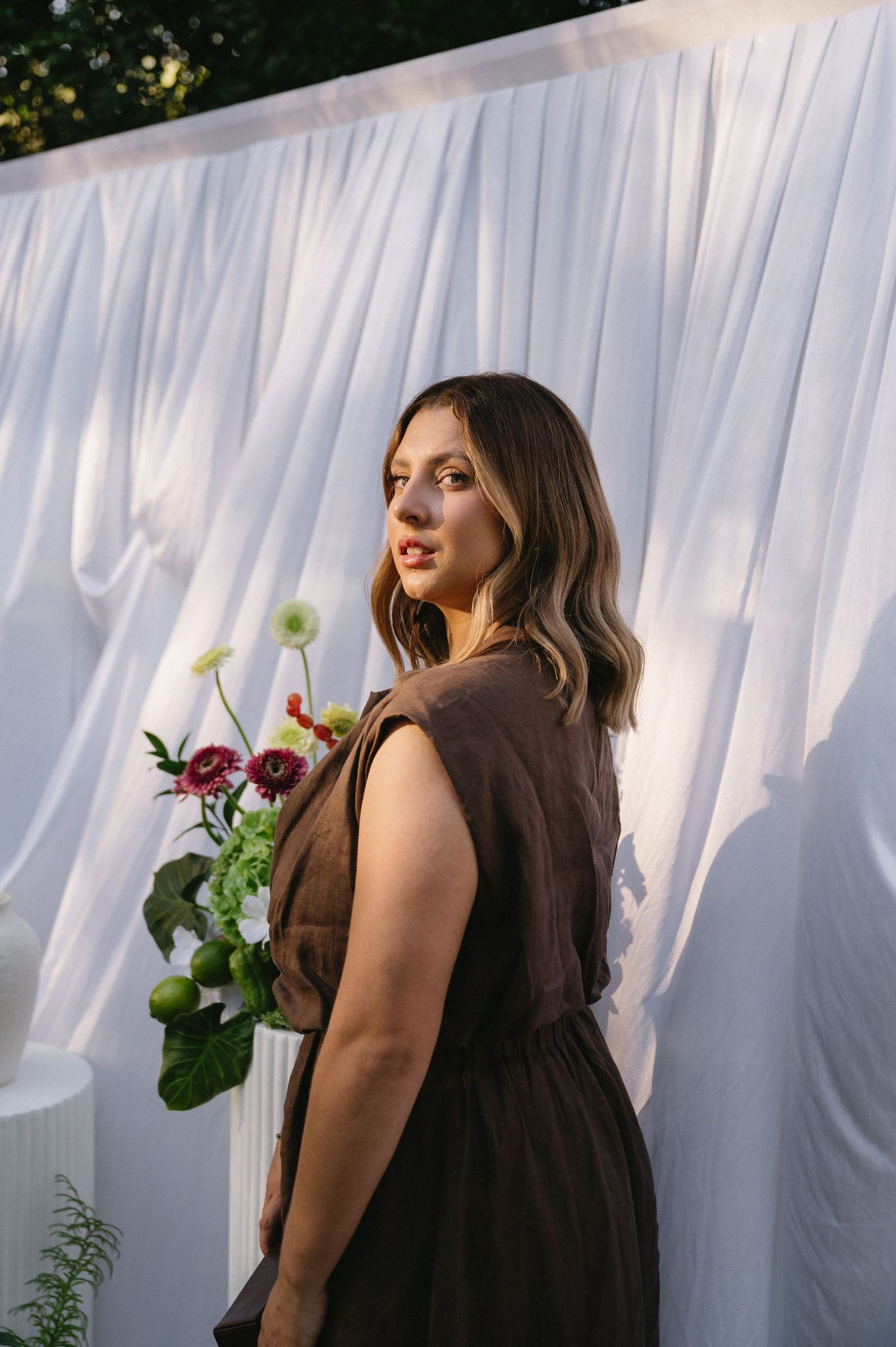 Woman in black dress poses against white curtain backdrop in outdoor photoshoot series.
