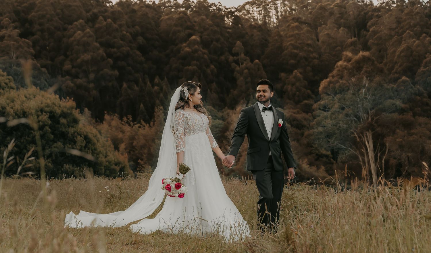 A romantic wedding couple stands in a golden field at sunset with trees in the background.