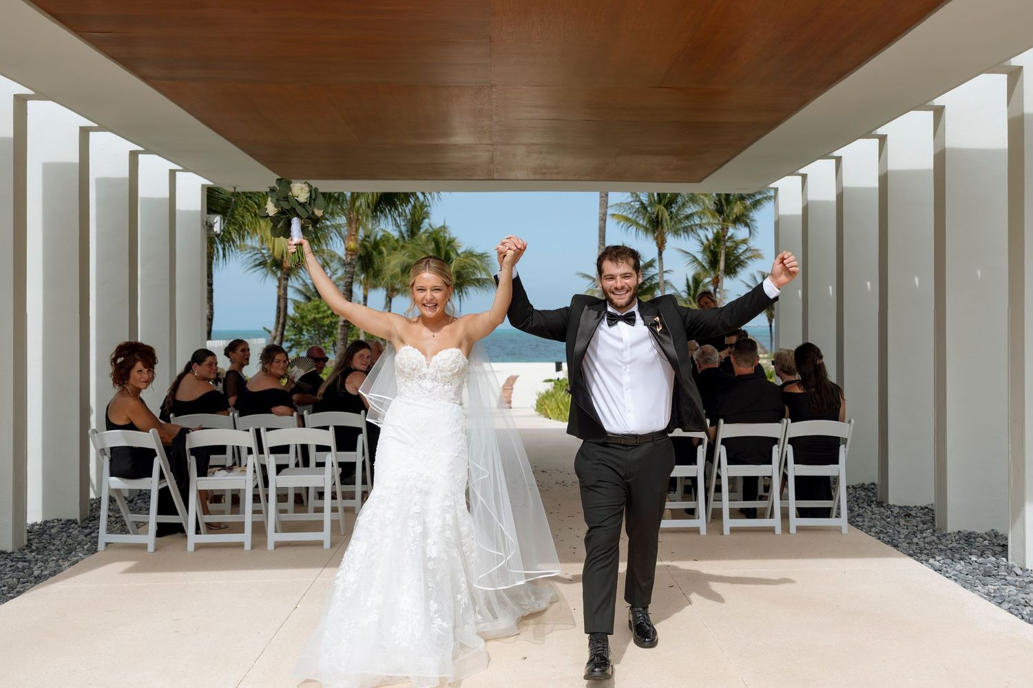 A joyful couple celebrates during their wedding ceremony at a modern beachside venue with white columns and palm trees.