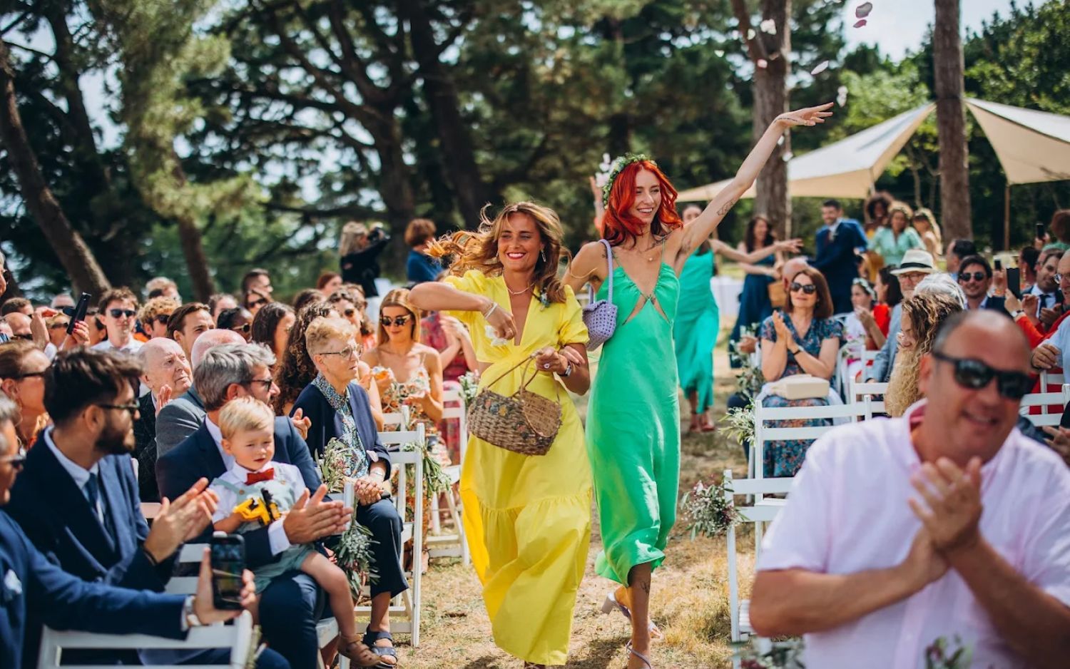 Wedding guests celebrate and cheer as bridesmaids in bright yellow and turquoise dresses walk down the outdoor aisle.