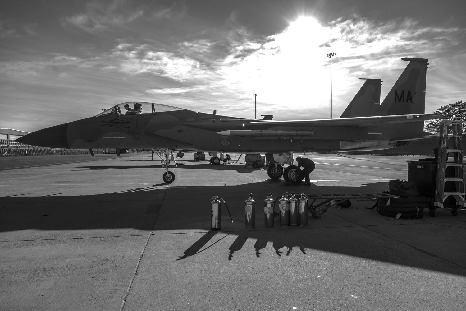 Military fighter jet silhouetted against dramatic sunset sky with ground crew shadows visible below.