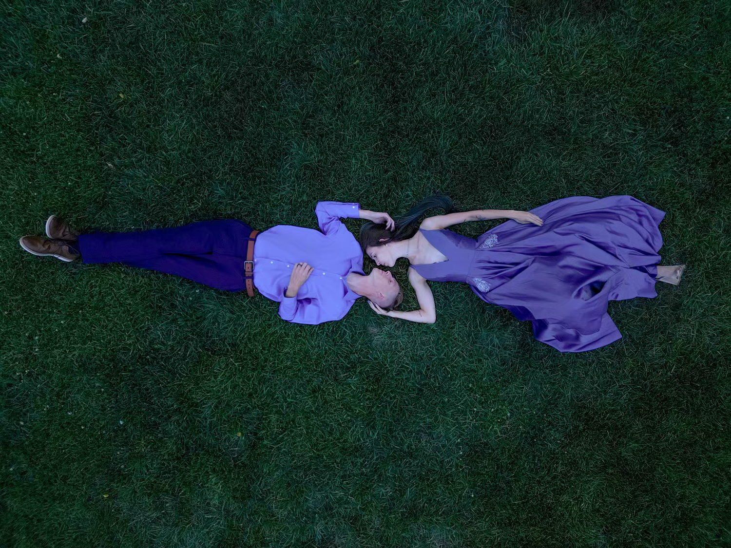 A couple in purple and blue lies on grass during a twilight photoshoot.