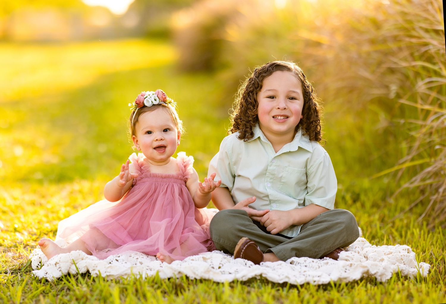 Children gather together outdoors smiling and sitting on a white blanket in warm sunlight enjoying a lovely day.