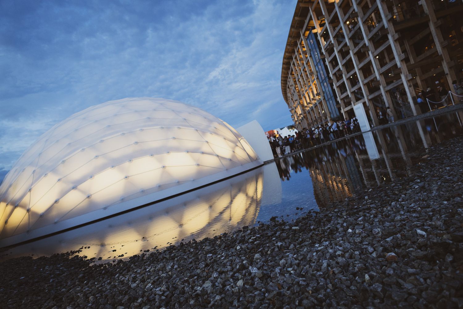 Illuminated dome structure reflected in water at dusk beside the woven wooden Portugal Pavilion exterior.