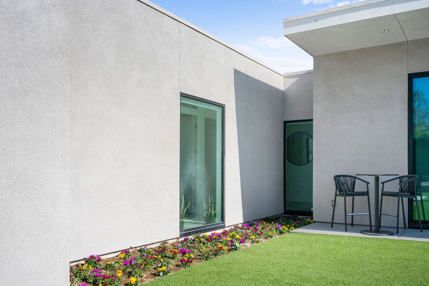 Modern stucco home exterior with green windows, lush lawn, colorful flower bed, and patio chairs under blue sky.