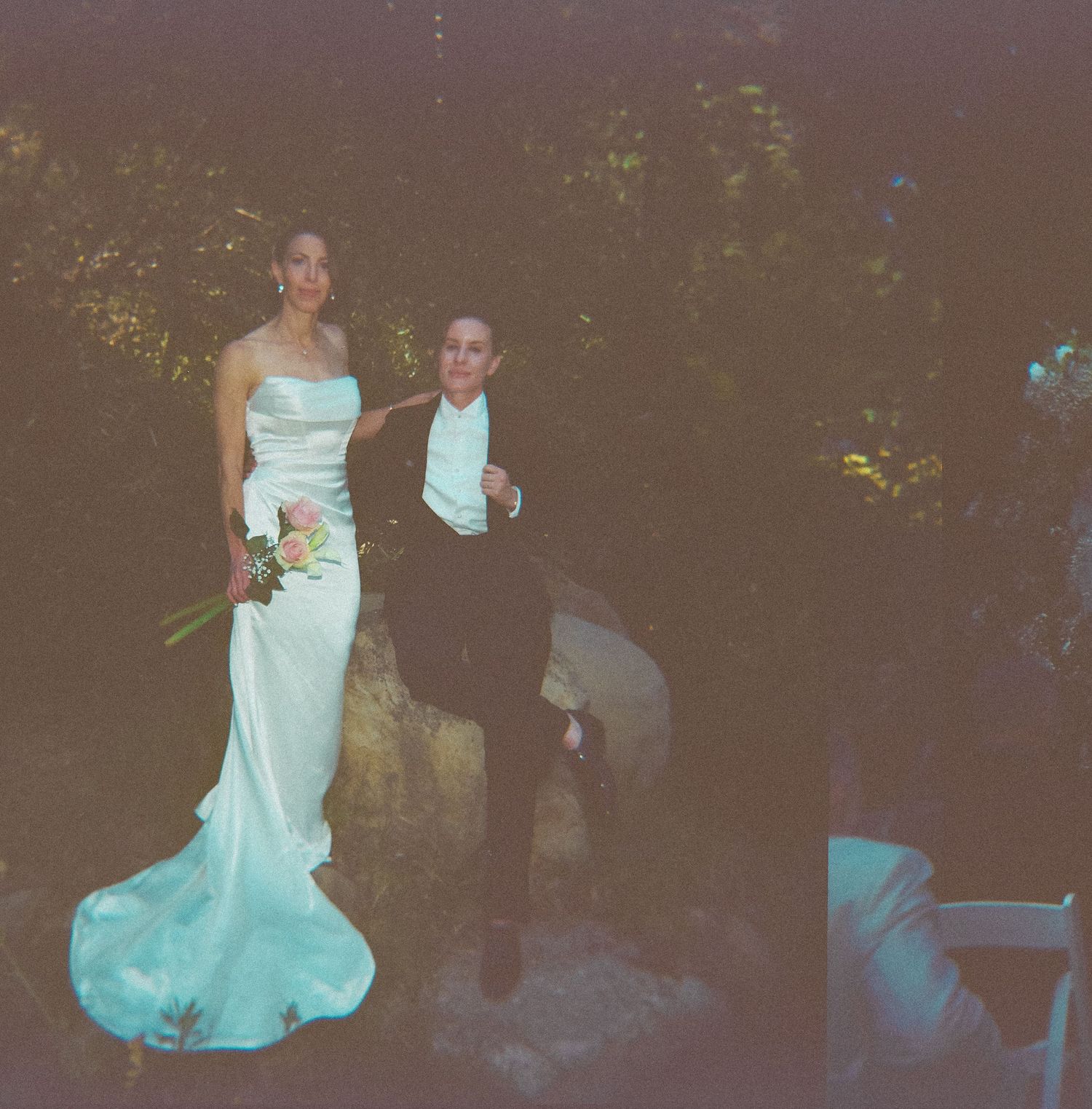 Bride in white gown holding bouquet stands with groom in black tuxedo outdoors at night during wedding ceremony.
