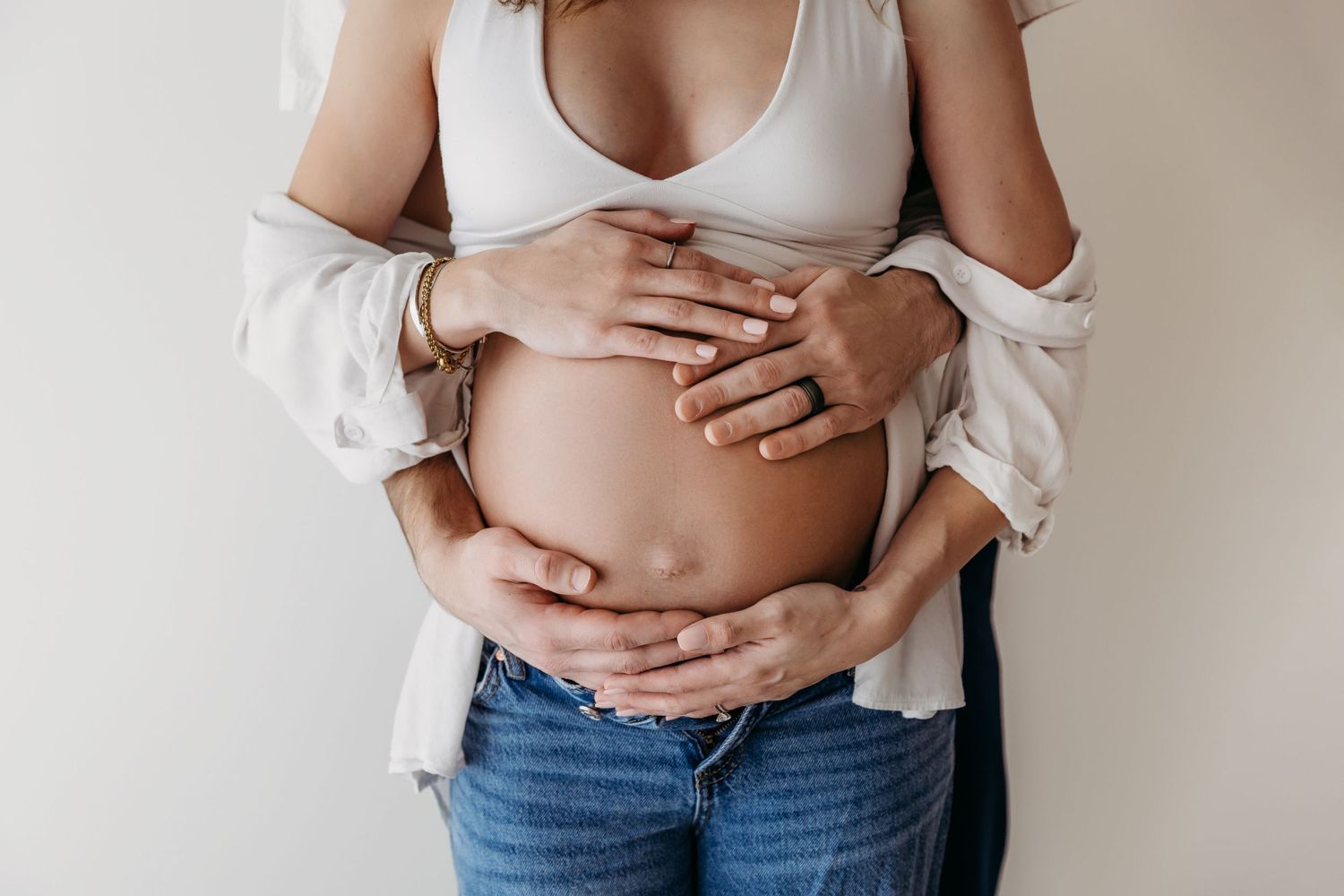 A pregnant woman in white top and jeans cradles her growing baby bump against a neutral background.
