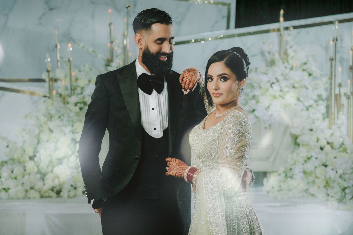 A couple in traditional Indian wedding attire stands in an elegant portico with a vintage green car in the background.