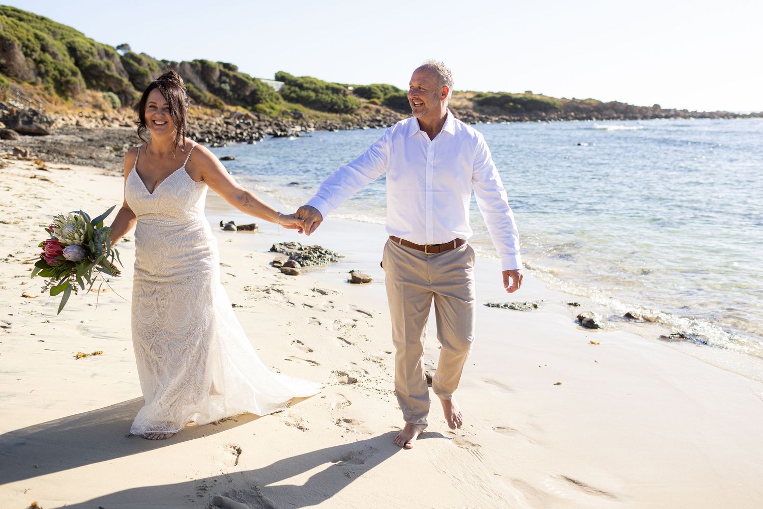 A couple in wedding attire embraces on a sunny rocky beach with ocean waves and coastal scenery in the background.